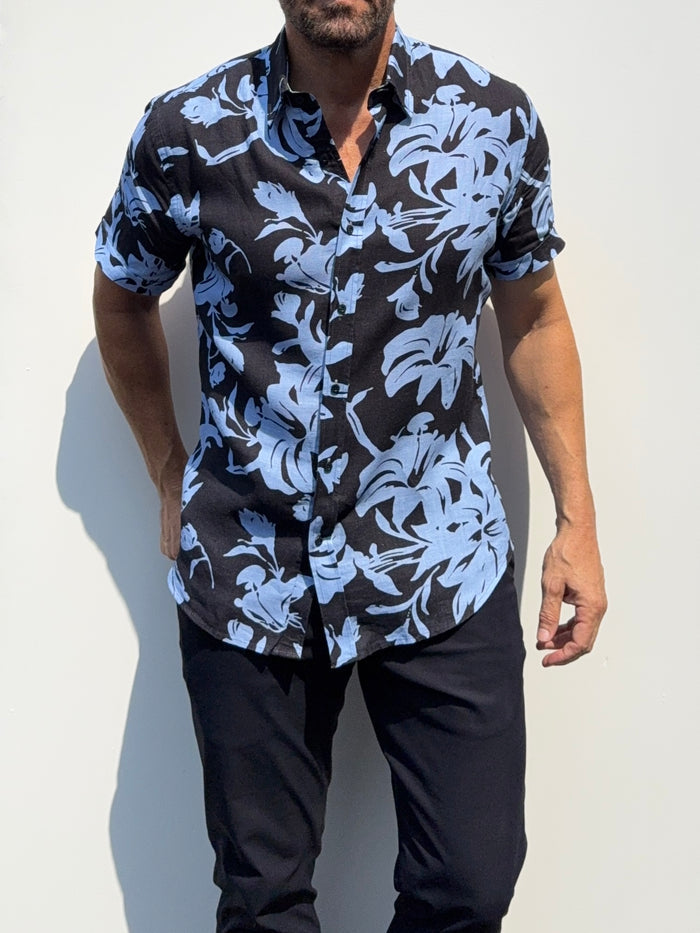 A man wearing a short-sleeve button-up shirt with a black base and a light blue tropical floral print, paired with dark pants, standing against a plain light background.