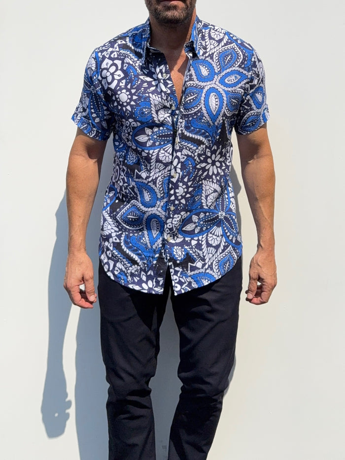 A man wearing a short-sleeve button-up shirt with a blue, white, and black paisley pattern, paired with dark pants, standing against a plain light background.