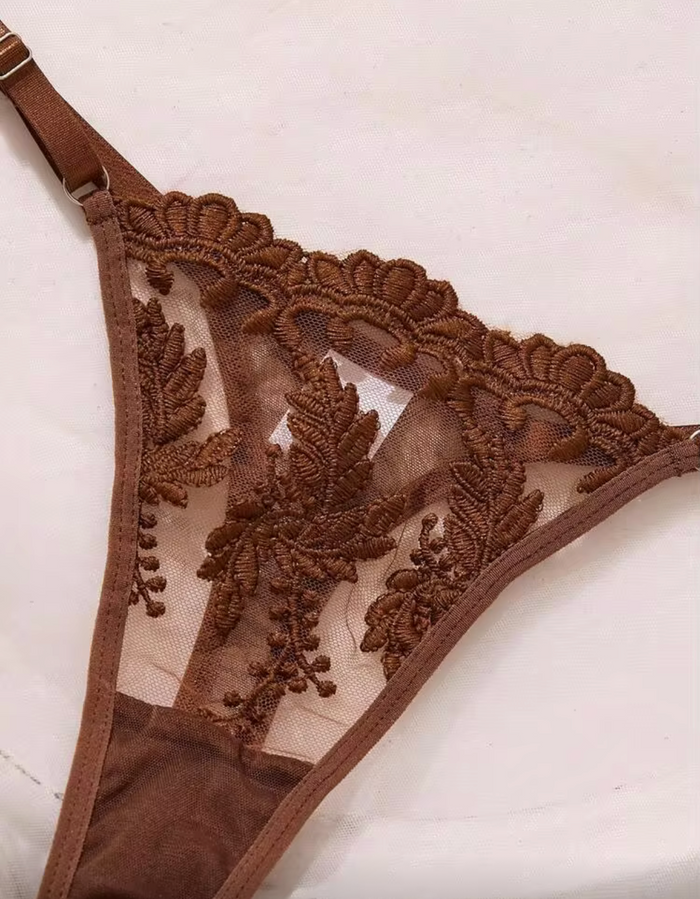 A close-up of a brown lace and mesh thong with floral embroidery, shown on a light beige background.