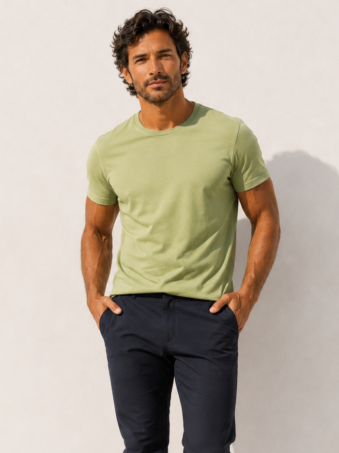 A man standing barefoot against a white wall, wearing a light sage green short-sleeve crewneck t-shirt and dark navy blue pants.
