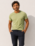 A man standing barefoot against a white wall, wearing a light sage green short-sleeve crewneck t-shirt and dark navy blue pants.