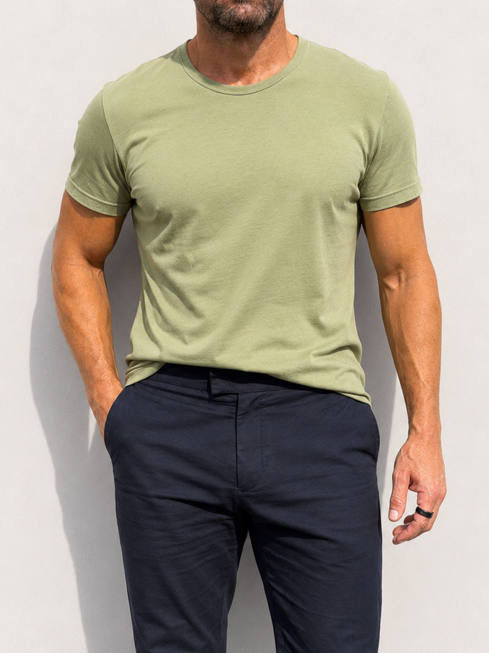 A man wearing a light olive green crew-neck t-shirt and dark navy blue trousers, standing against a plain light gray wall.