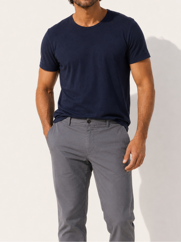 A man wearing a navy blue crewneck t-shirt and gray trousers, standing against a plain off-white wall with a soft shadow.