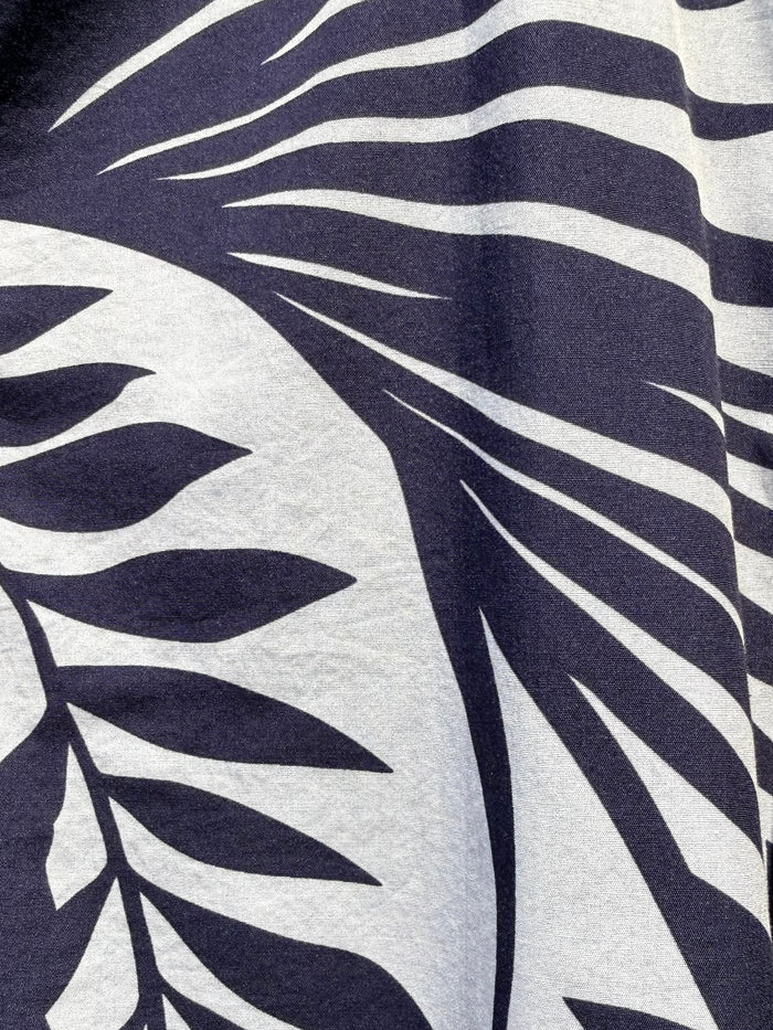 Close-up of a tropical leaf print fabric with dark navy blue palm fronds on a white background, showing part of a buttoned garment.