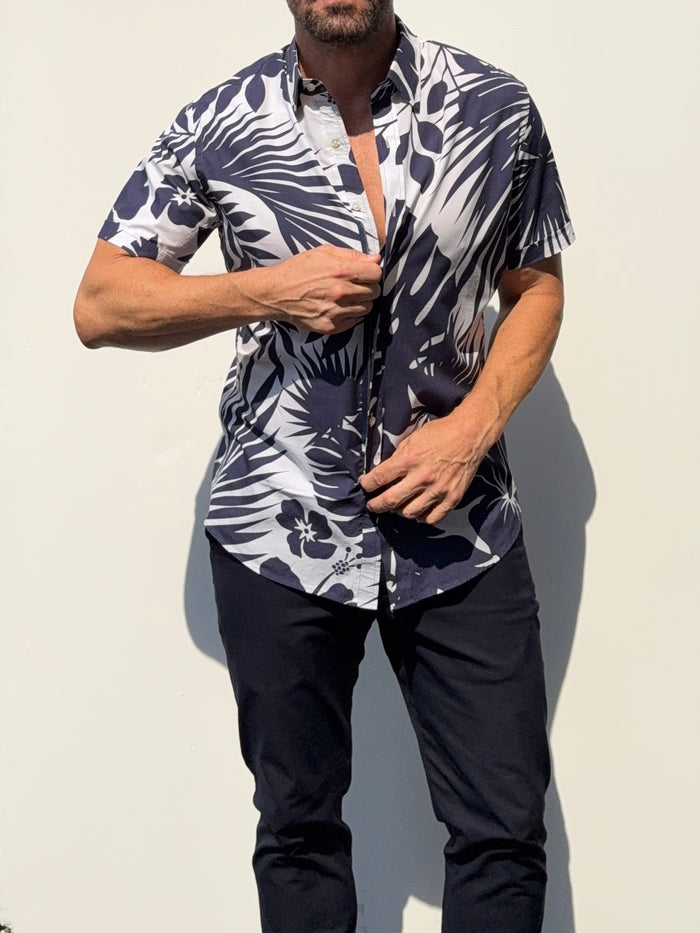 A man wearing a short-sleeve button-up shirt with a bold navy blue and white tropical leaf print, paired with dark pants, standing against a plain light background.