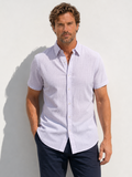 Man wearing a light purple short-sleeve shirt and dark pants against a white wall.