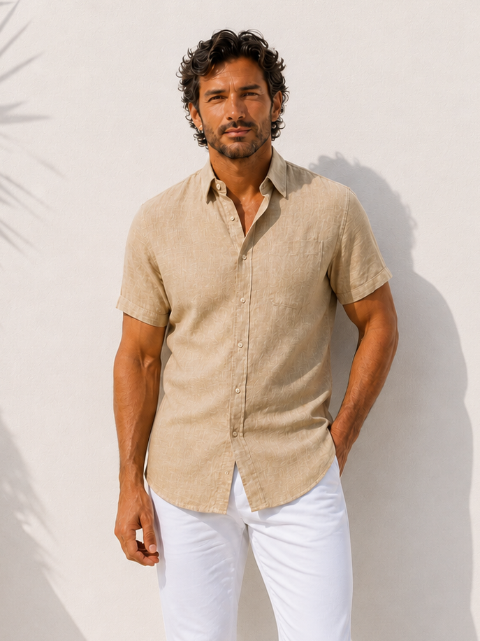 Man wearing a beige shirt and white pants standing against a white wall with a plant to the side.