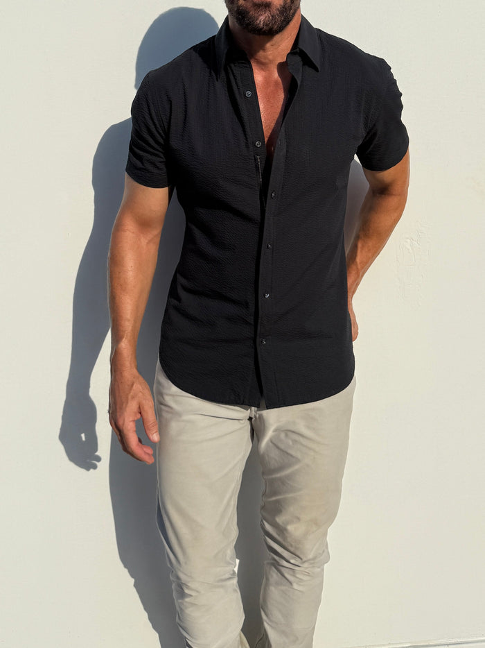 Man wearing a black short-sleeve shirt and light gray pants against a white background