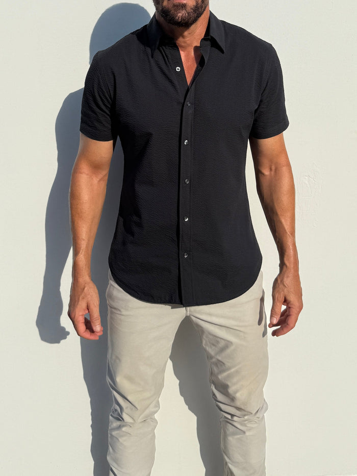Man wearing a black short-sleeve shirt and light gray pants against a white background