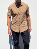 A man wearing a short-sleeve button-up shirt in a warm tan or camel color, paired with dark navy pants, standing against a plain white background.