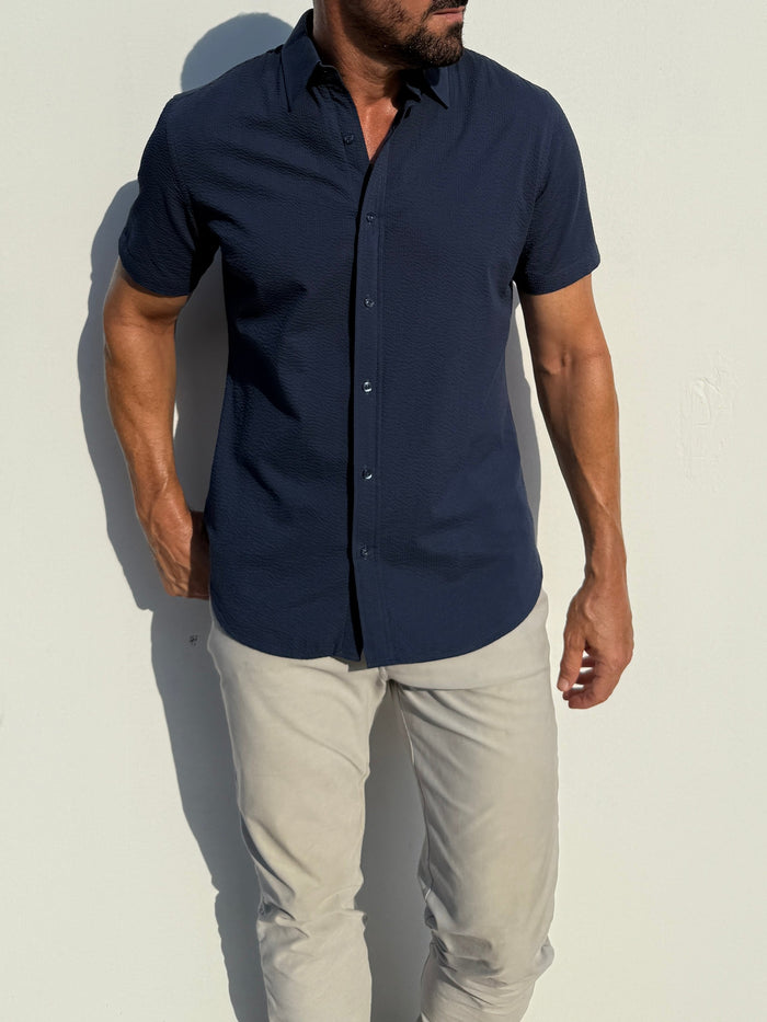 Man wearing a navy blue short-sleeve shirt and light gray pants against a white background