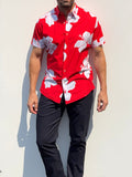 A man wearing a red short-sleeve button-up shirt with a white hibiscus floral print, paired with dark pants, standing against a plain white wall.