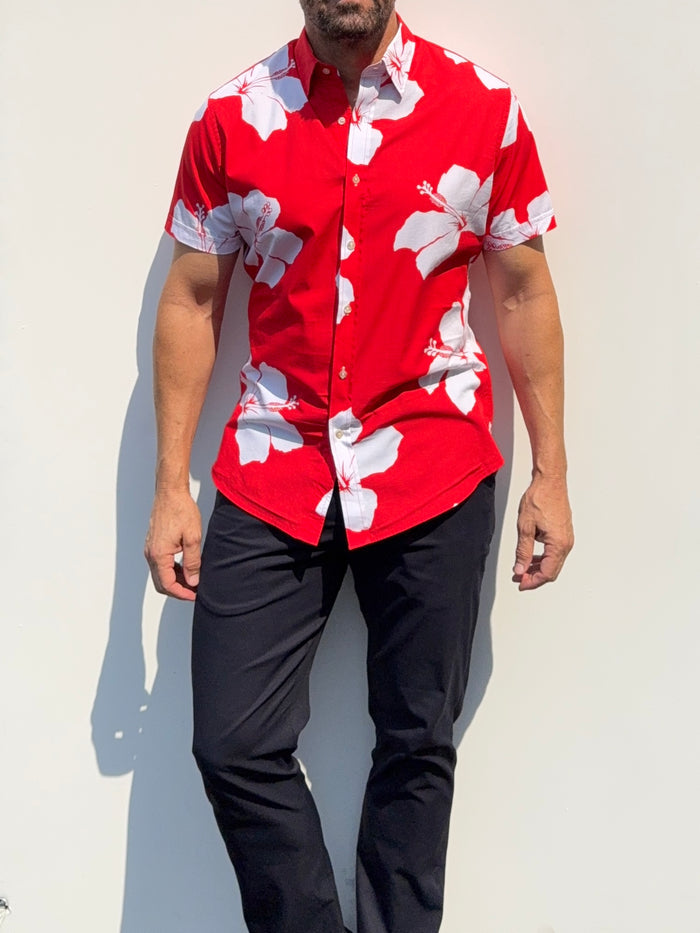 A man wearing a red short-sleeve button-up shirt with a white hibiscus floral print, paired with dark pants, standing against a plain white wall.